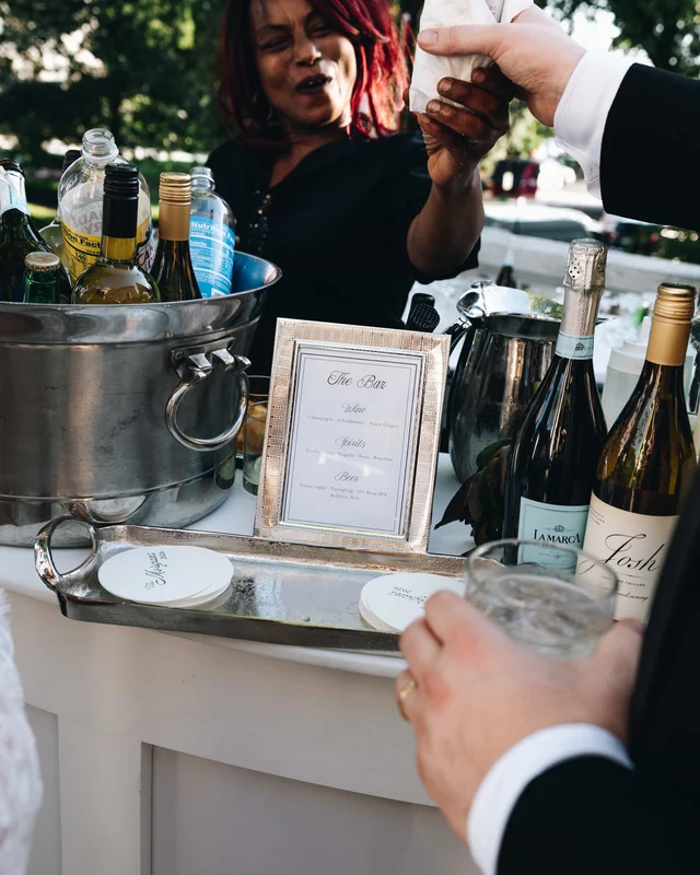 A bartender reaches across the iced bottle tub while a guest's glass waits beside the framed bar menu