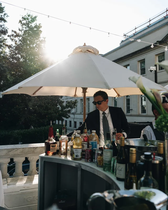 The bartender works beneath a white umbrella at golden hour, bottles crowding the curved bar