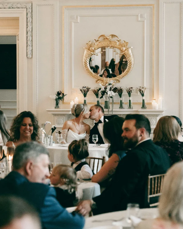 The couple steals a kiss at the sweetheart table beneath a gilt mirror and a mantel of calla lilies