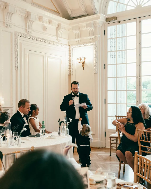 The best man reads his toast into the microphone while a toddler tugs at his jacket
