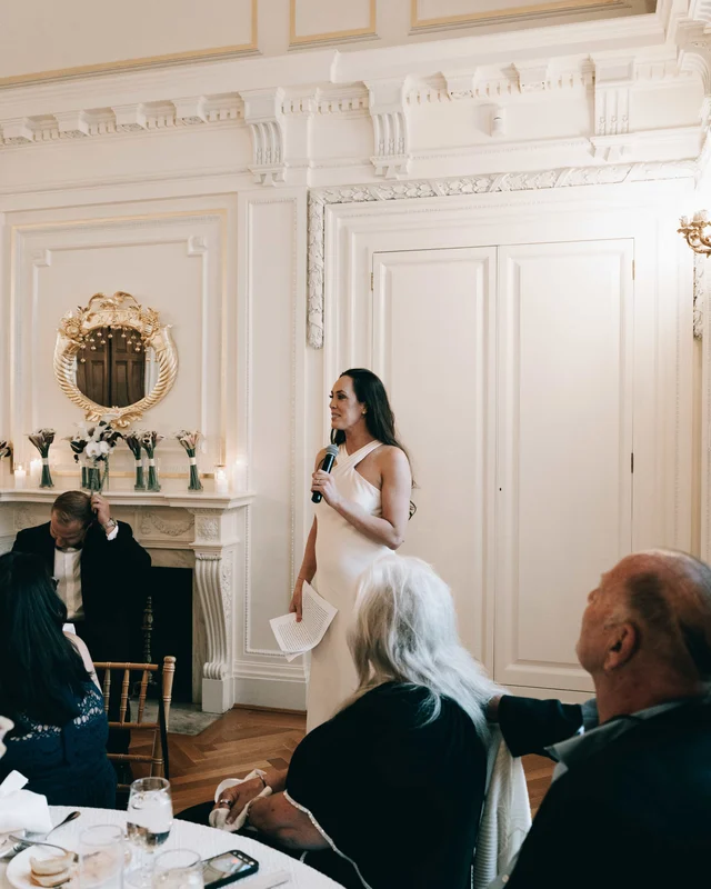 A dark-haired bridesmaid delivers her toast, speech in one hand and microphone in the other