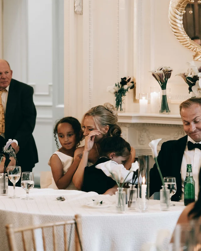 The bride wipes away a tear at the head table, a small child pulled close against her