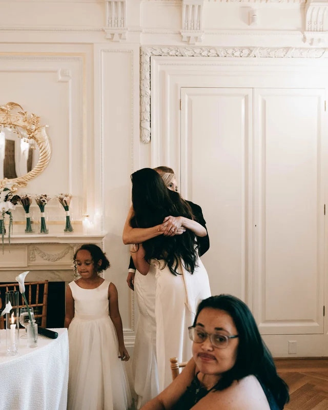 The bride folds the toast-giver into a hug by the mantel while the flower girl looks on