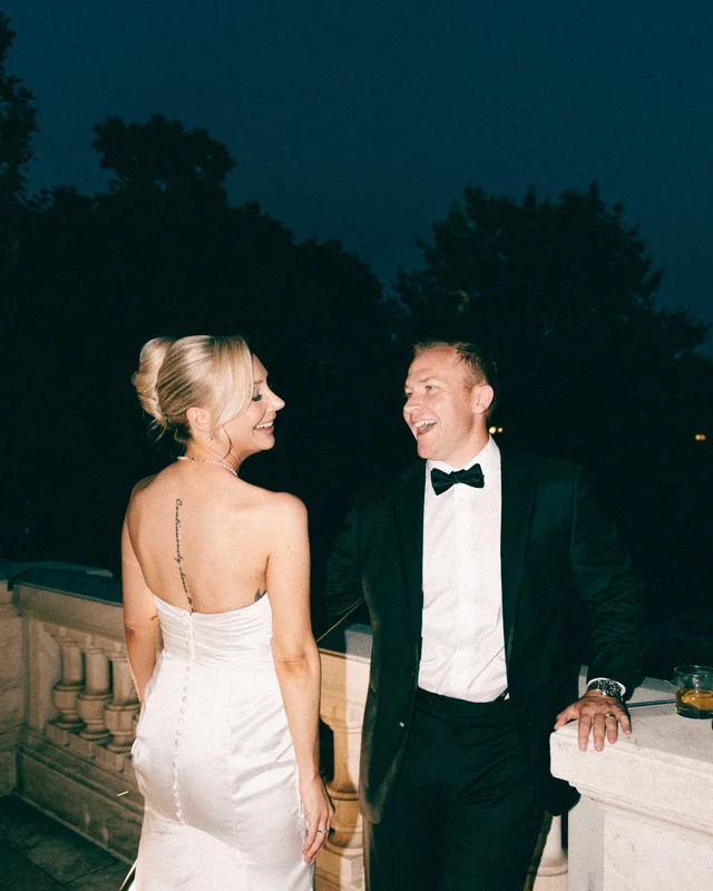 Night flash catches the couple laughing at the balustrade, his hand resting near a glass of whiskey