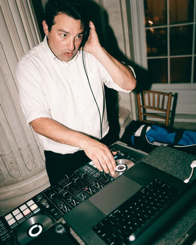 The DJ cups his headphones with one hand and rides the mixer with the other