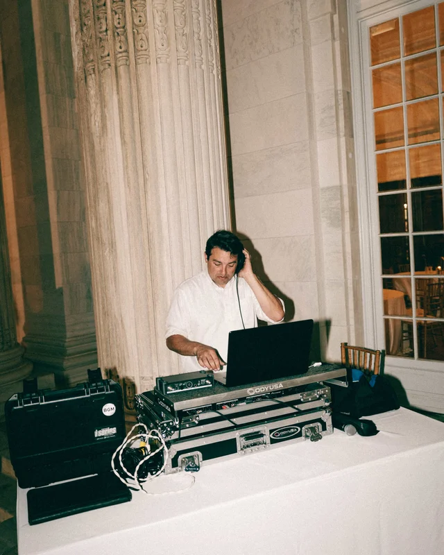 The DJ stands behind his road-cased rig beside a marble column, cables coiling across the linen