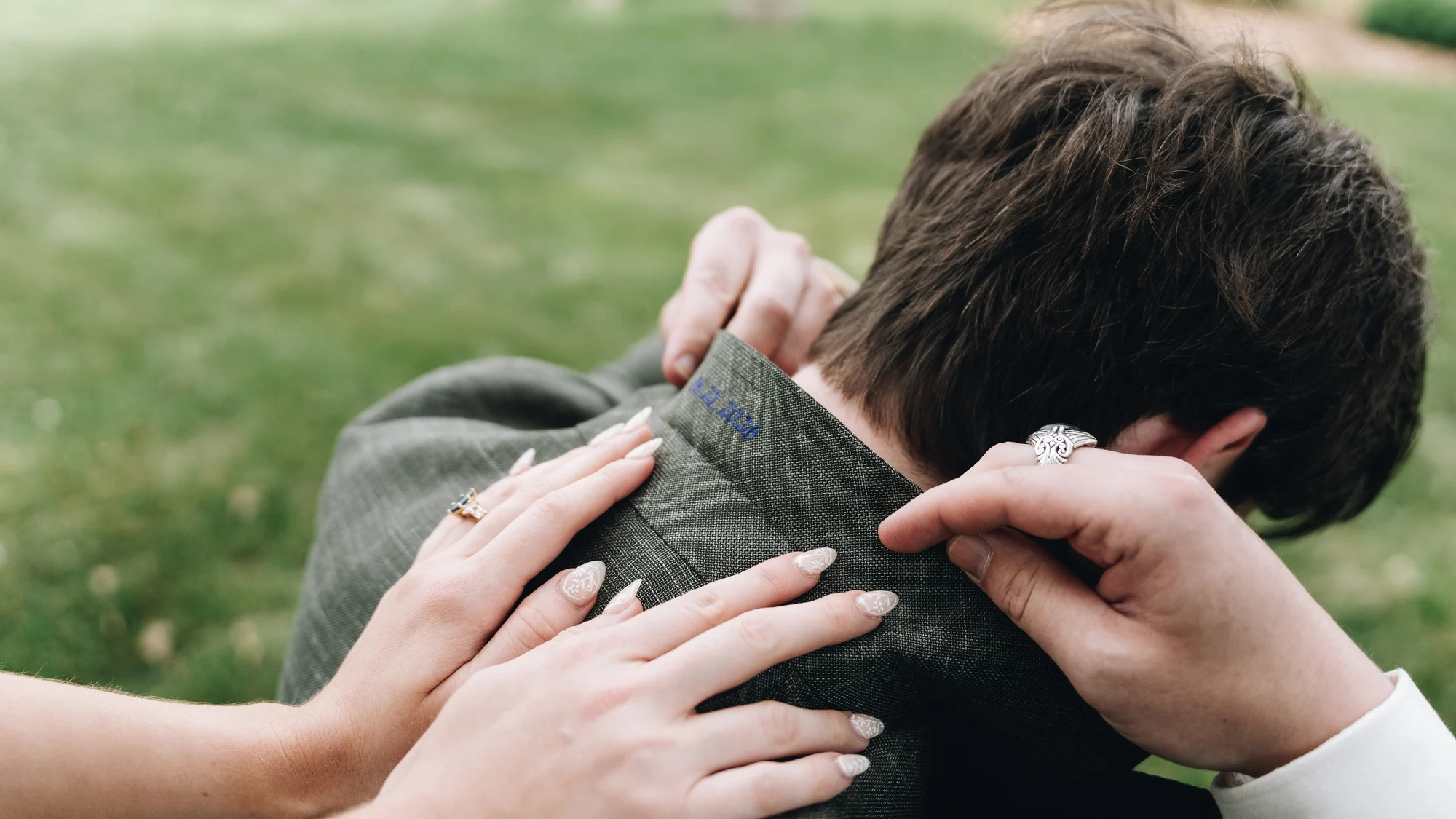 Close on the bride's hands folding back the groom's jacket collar, where he had had their wedding date embroidered in blue thread