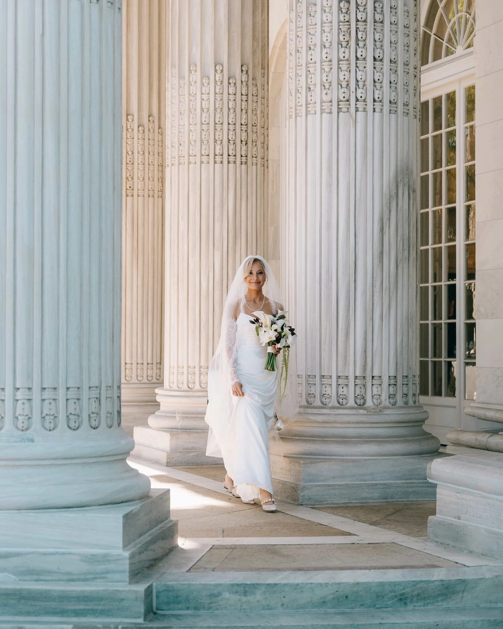The bride carrying her callas between the towering fluted columns, her veil back and a smile already going