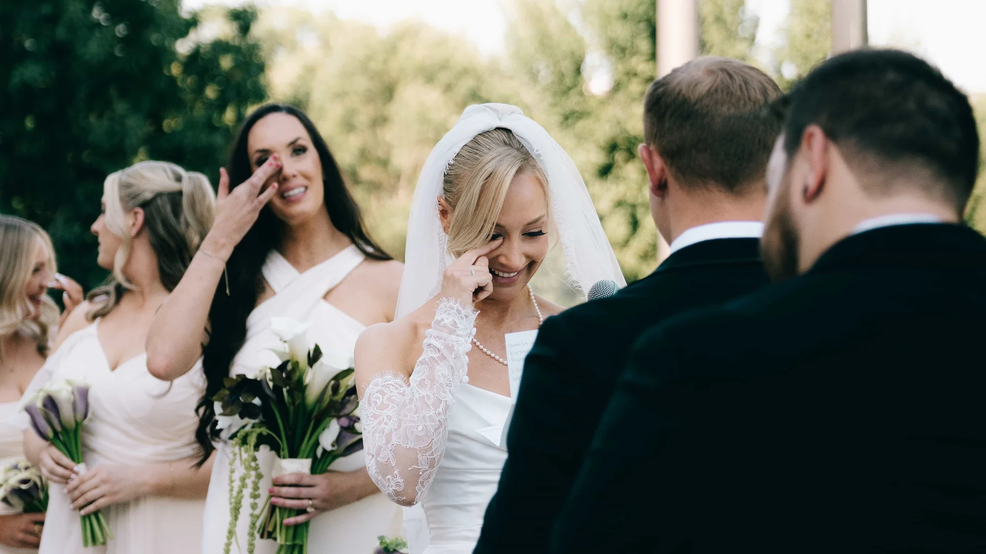 The bride laughing through a tear during the vows, a bridesmaid behind her wiping her own