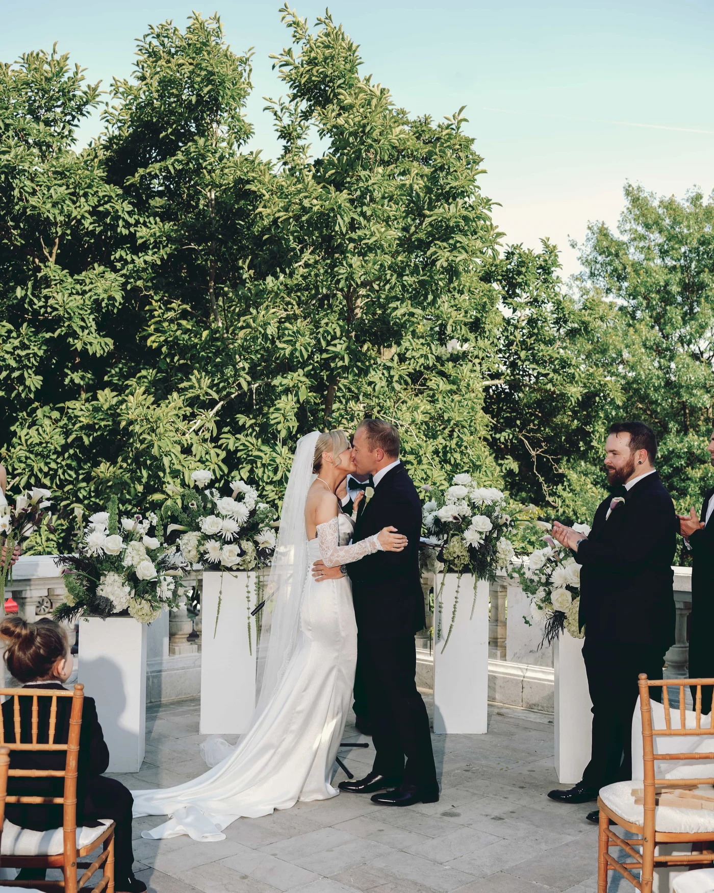 The kiss under the trees between pedestals of white flowers, a small boy watching from his chair