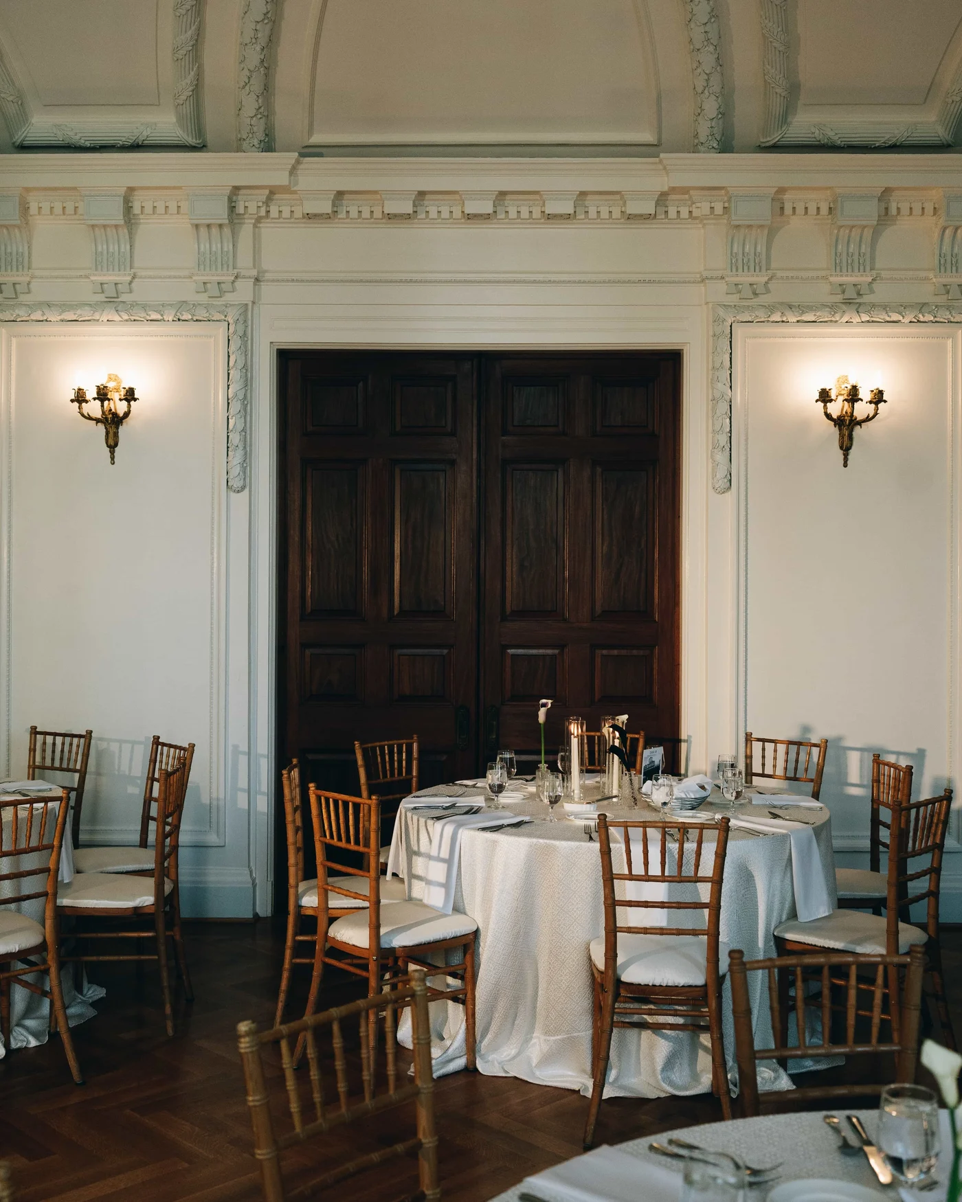 A round table set in white before the dark double doors, brass sconces lit on either side
