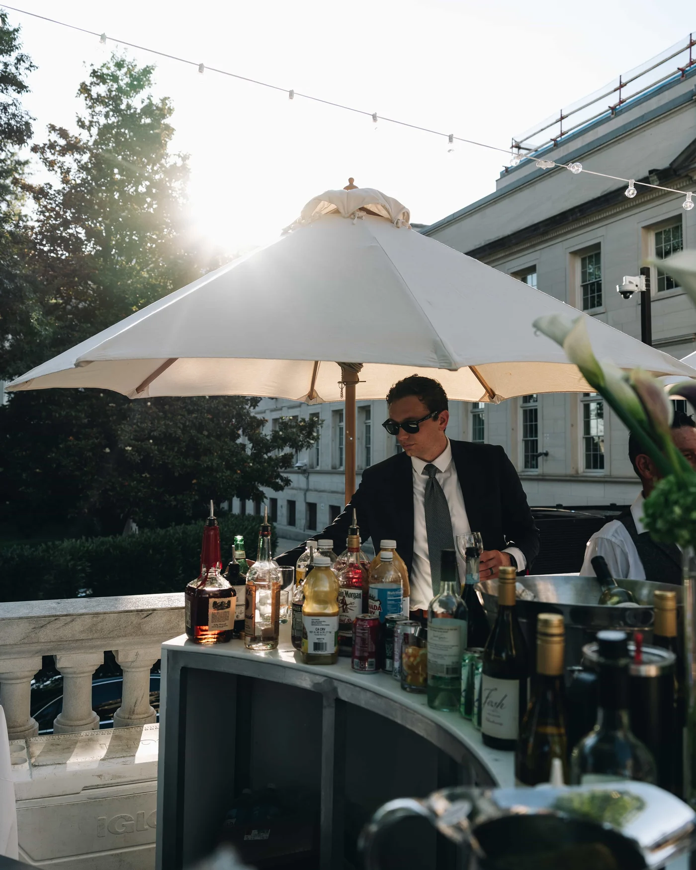 A bartender in sunglasses working under a white umbrella, bottles crowded in front of him and the sun flaring behind