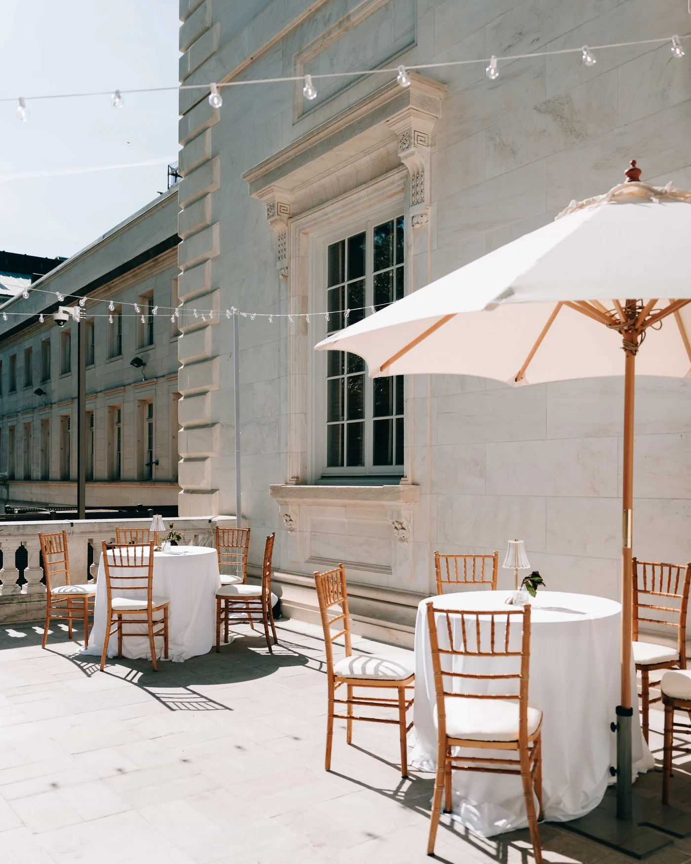 Cocktail tables and gold chairs under string lights and a white umbrella, set against the marble facade