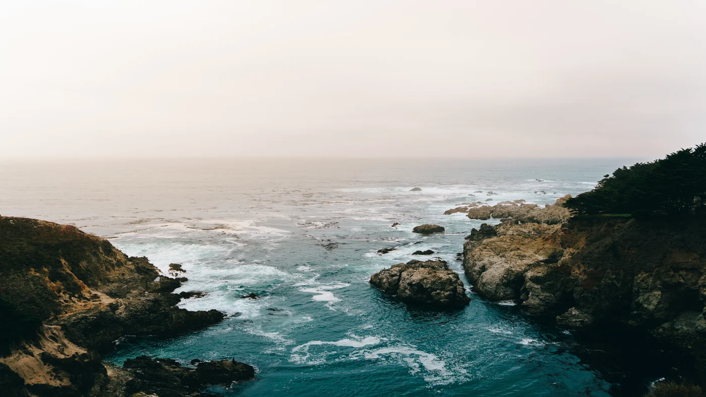 Surf churning around rocky islets in a cove, fog washing the horizon pale