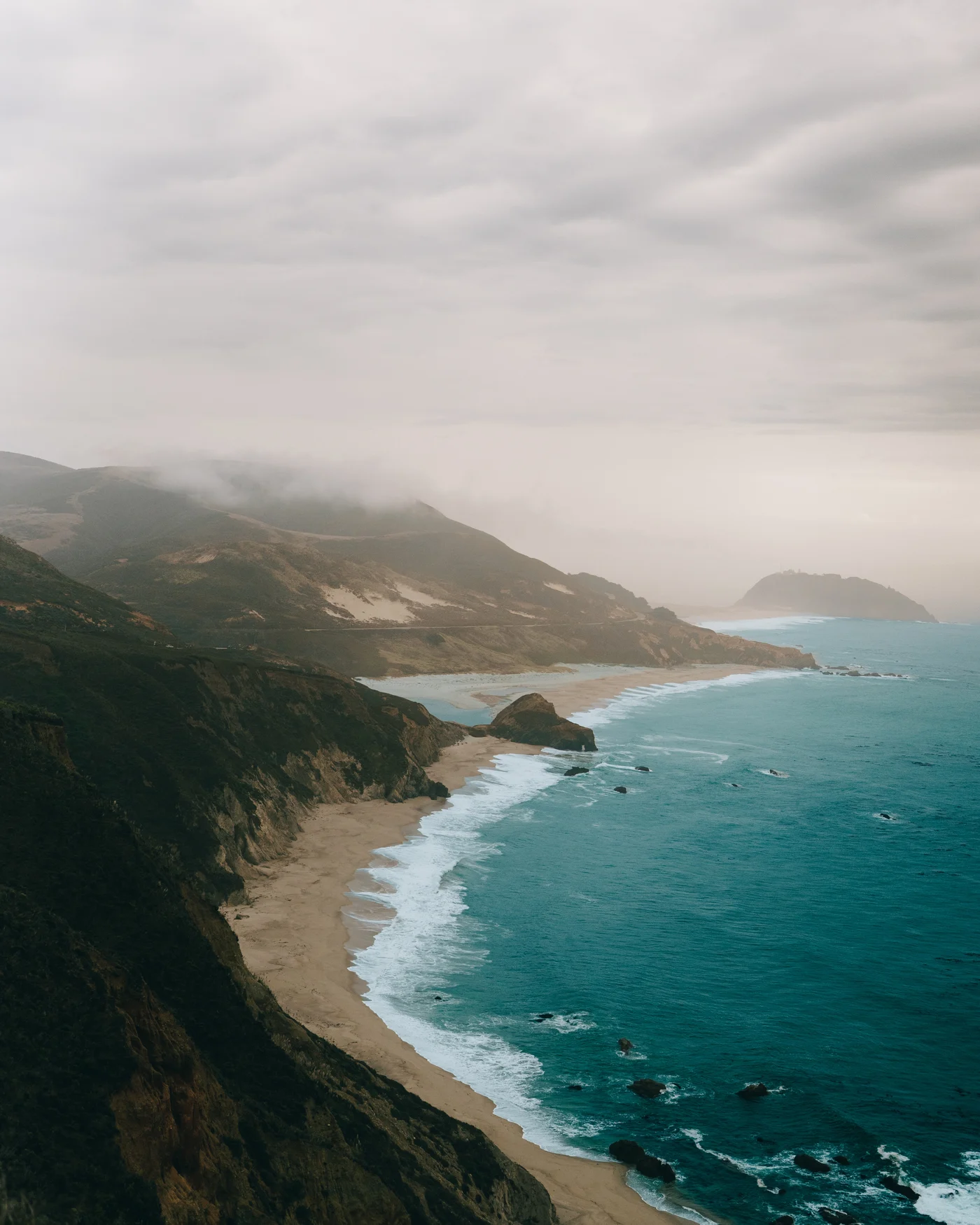 A crescent of sand curving beneath coastal bluffs, surf rolling in under low cloud