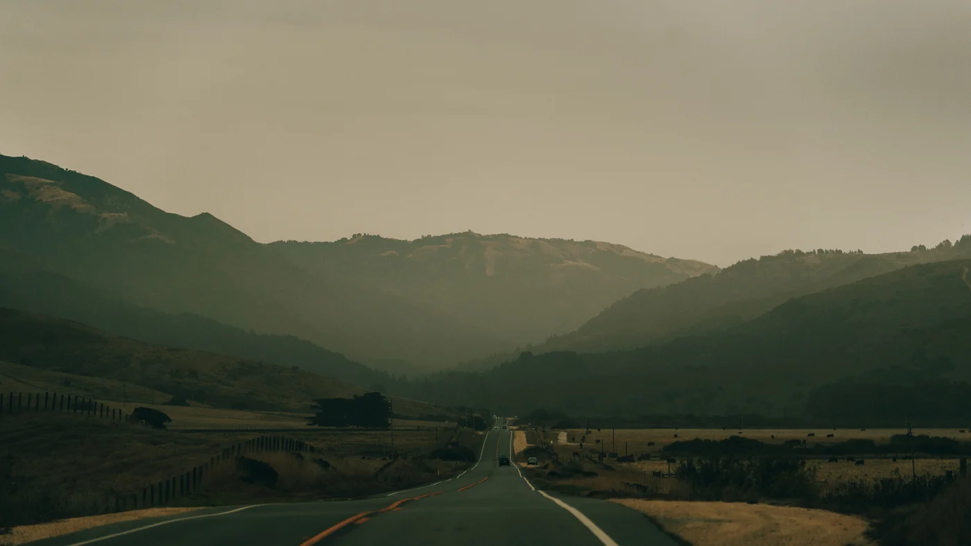 A lone car far ahead on a straight highway, hazy ridgelines stacked beyond