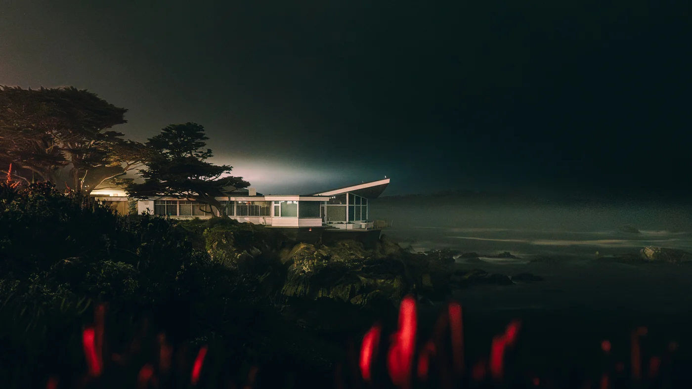 A midcentury glass house glowing at night above the rocks, red blooms blurred in the foreground