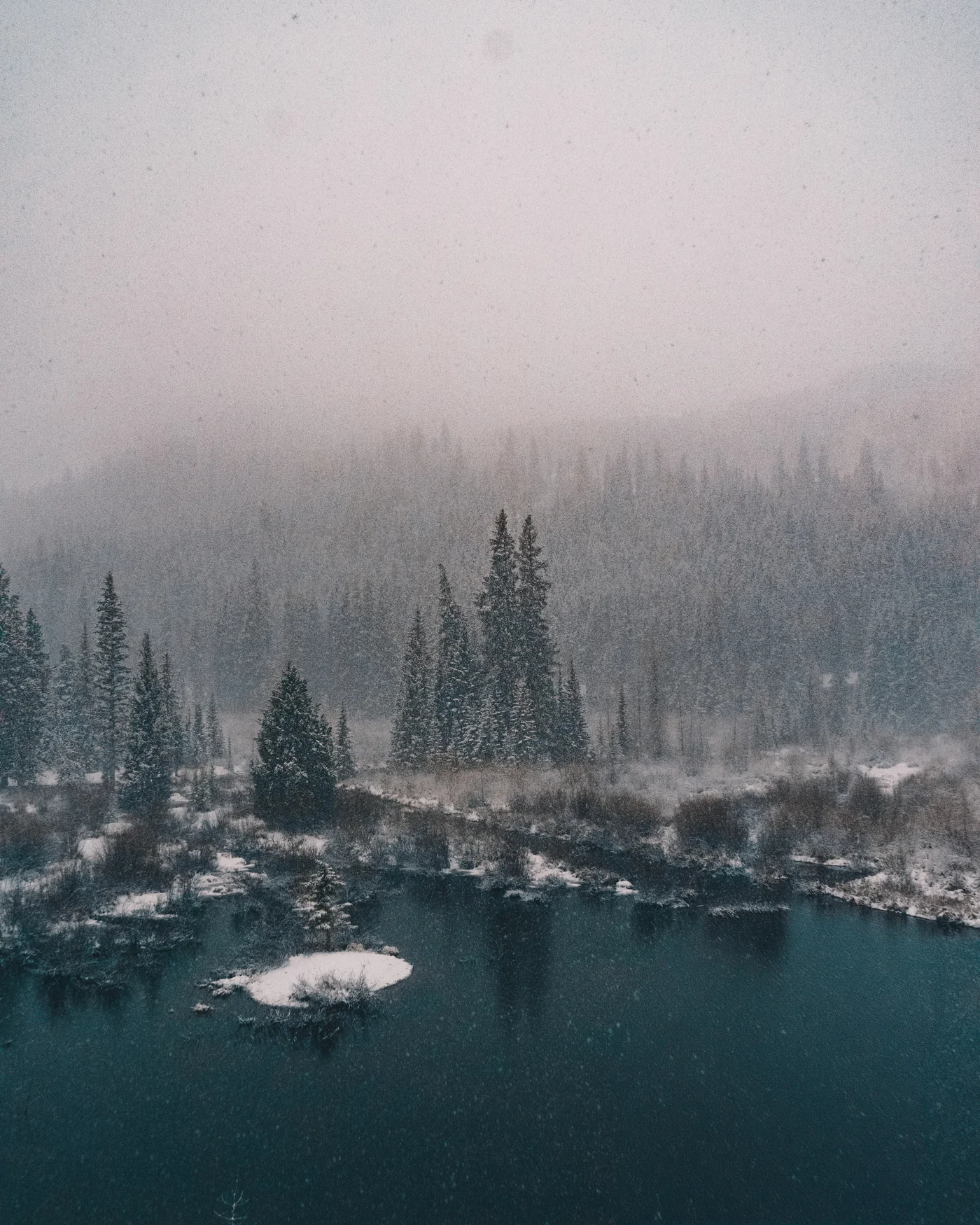 Snow falling over a dark river bend hemmed in by foggy pines