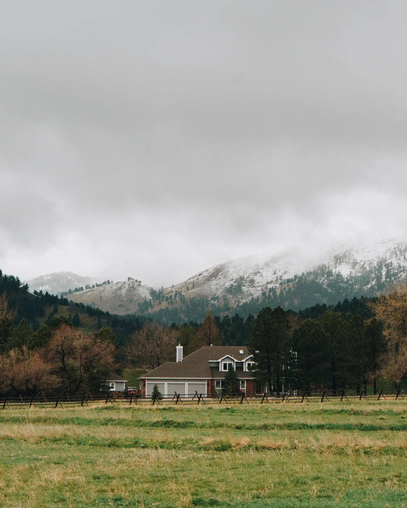 A ranch house tucked beneath snow dusted foothills, a rail fence running past