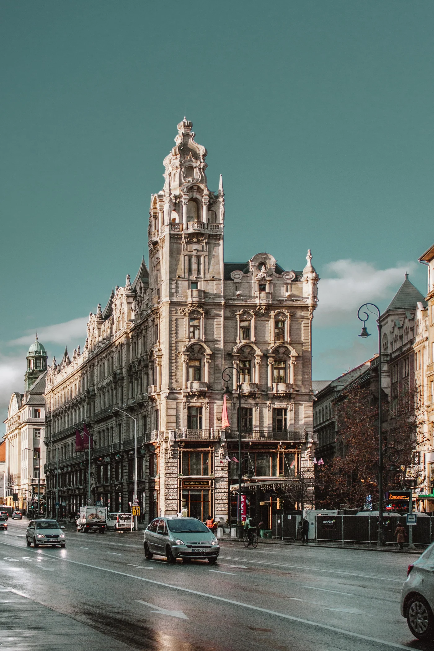 An ornate stone palace towering over a wet Budapest street, cars passing below