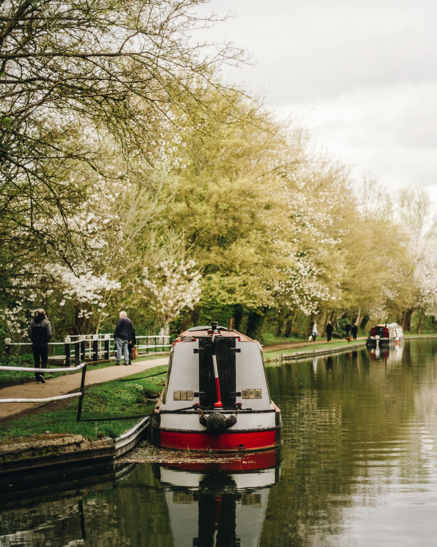 A red and white narrowboat moored on a canal beneath spring blossoms, walkers on the towpath