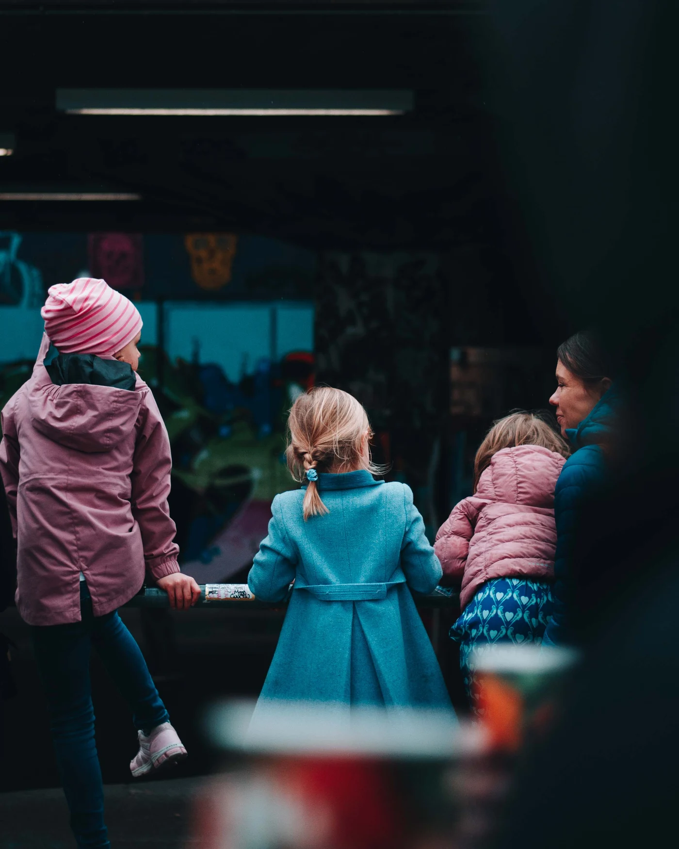 Three children in pink and blue coats gather at a rail beside a woman