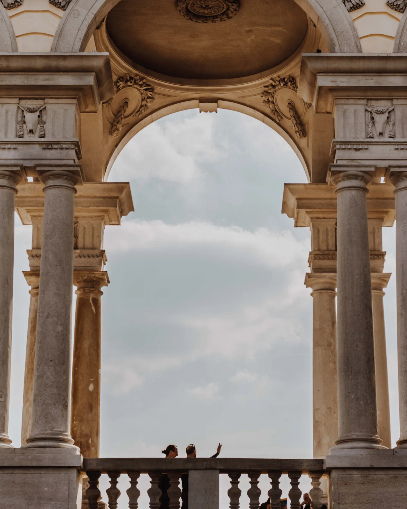 Two figures wave from a balustrade beneath a grand arched colonnade, clouds filling the arch
