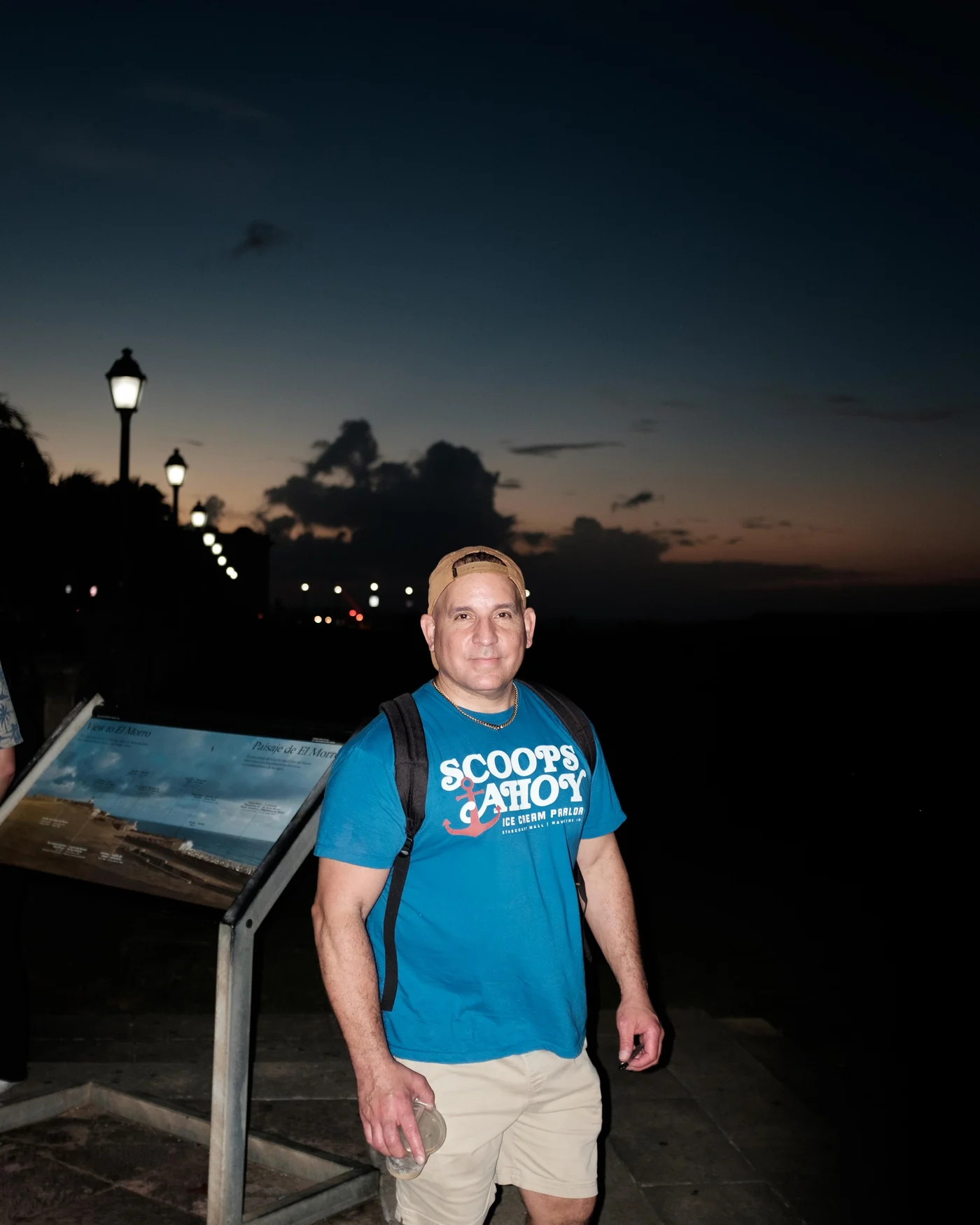 A man in a blue Scoops Ahoy shirt smiles at dusk, lampposts glowing down the promenade