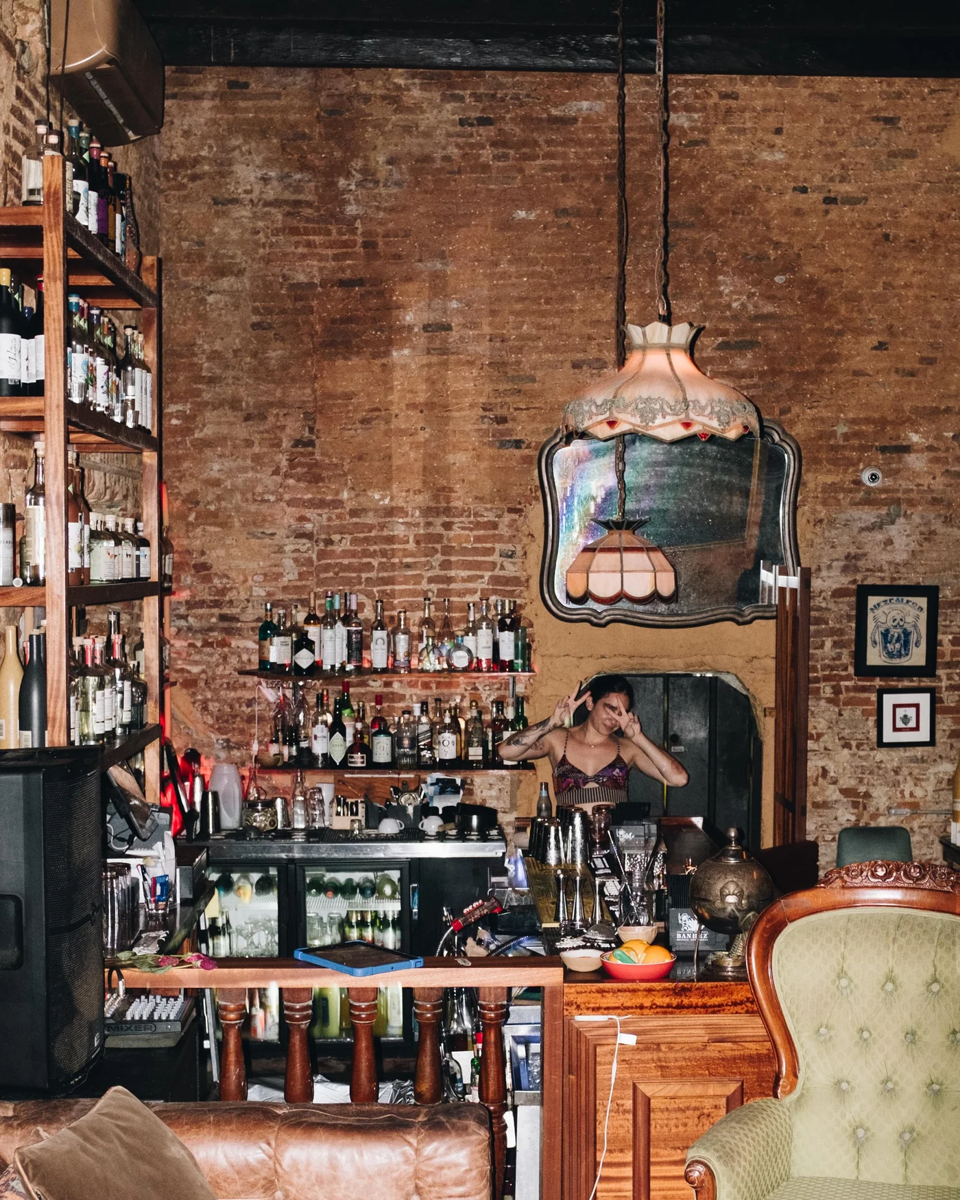 A bartender flashes peace signs beneath a stained glass lamp in a brick walled bar