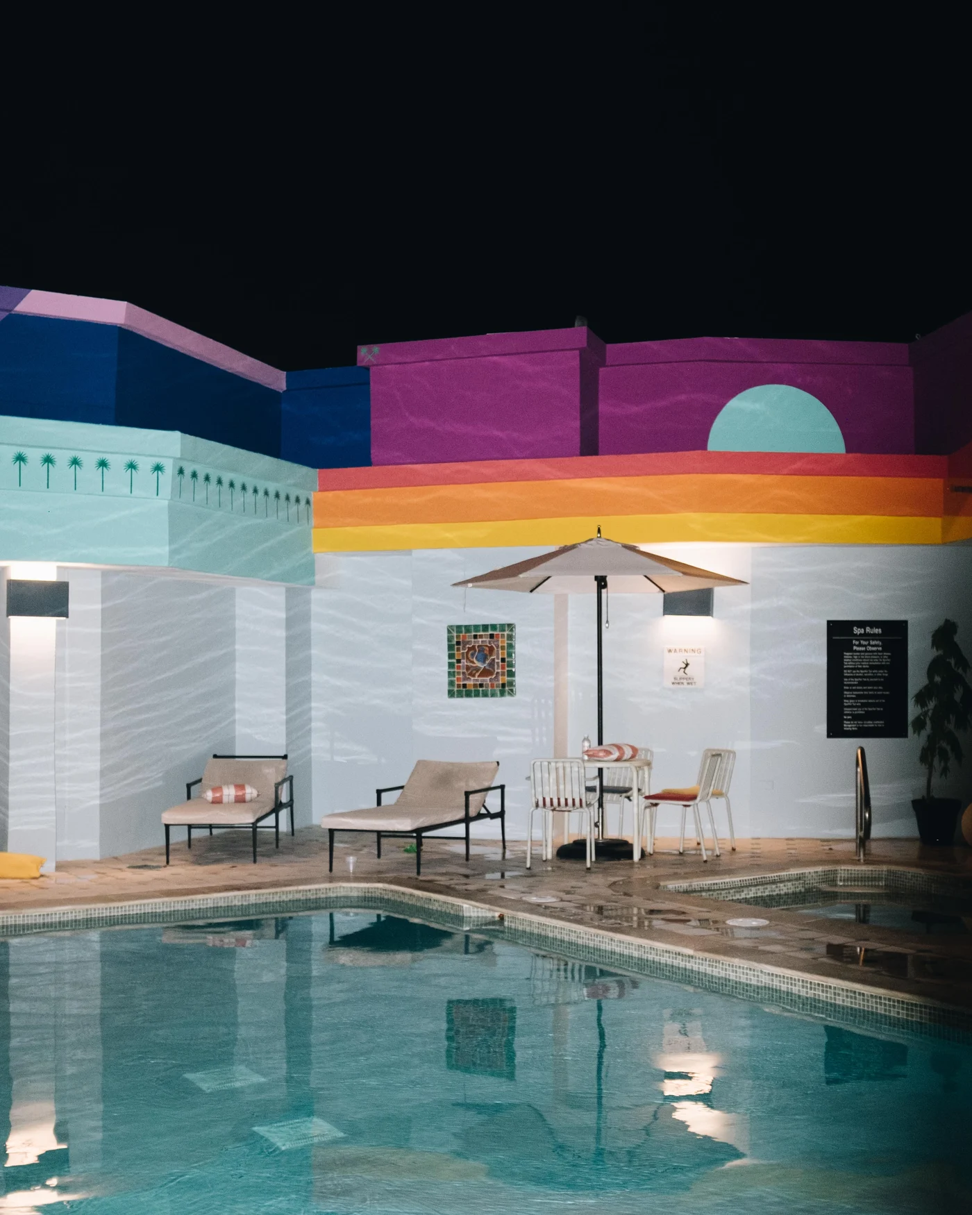 A rooftop pool at night, its walls painted in bands of mint, purple, and orange