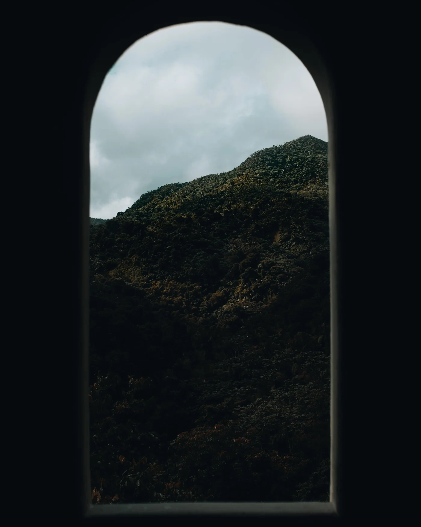 A green mountainside framed through a dark arched window, clouds resting on the ridge
