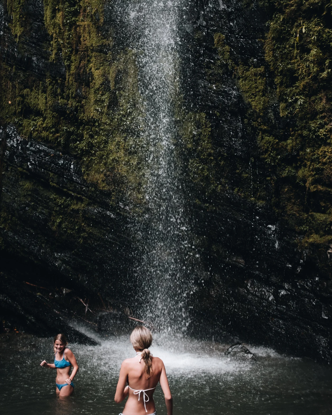 Two swimmers wading beneath a tall waterfall spilling down a mossy rock face