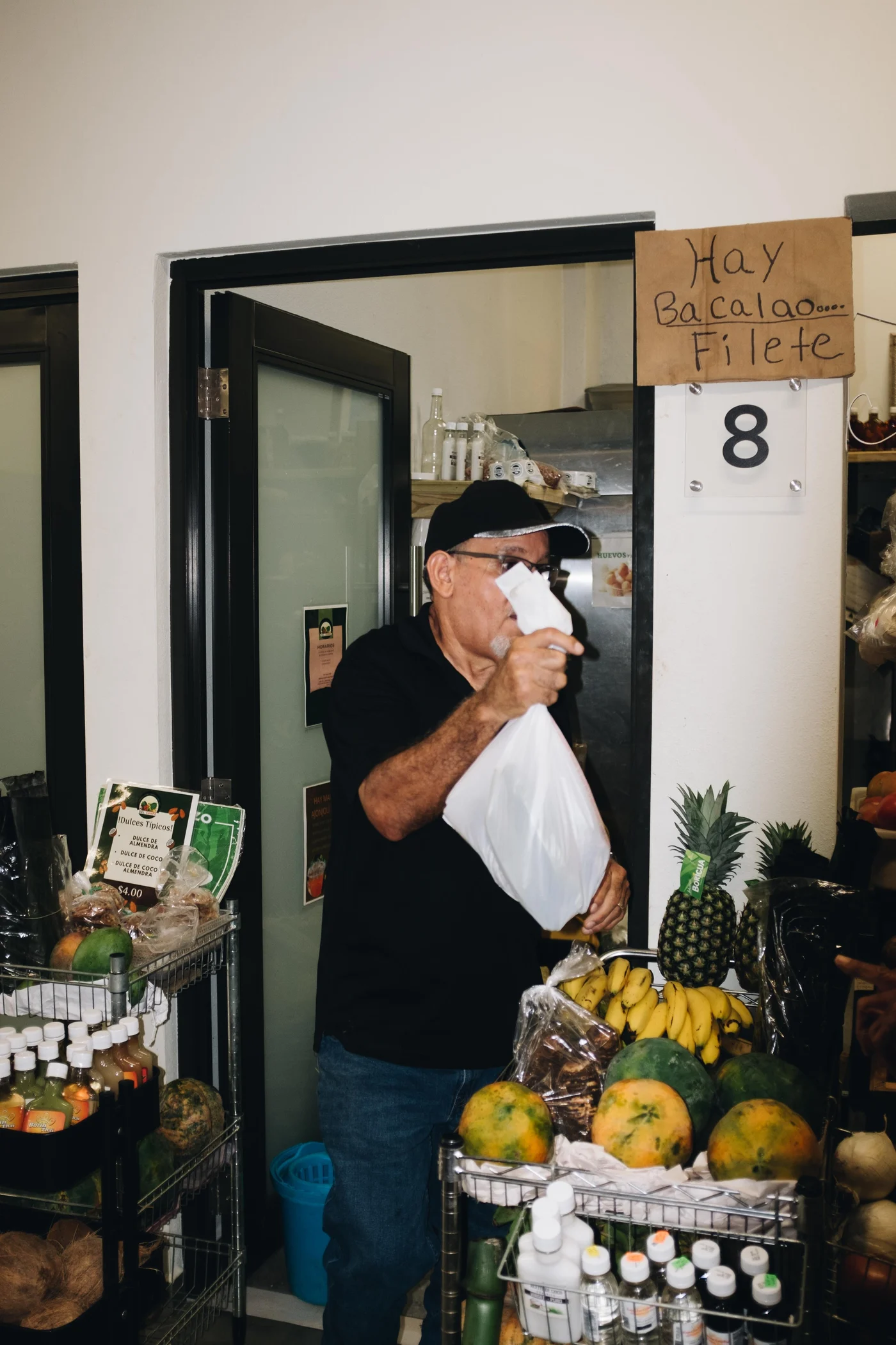 A grocer carries a white bag past bananas and a pineapple, a handwritten Hay Bacalao sign overhead