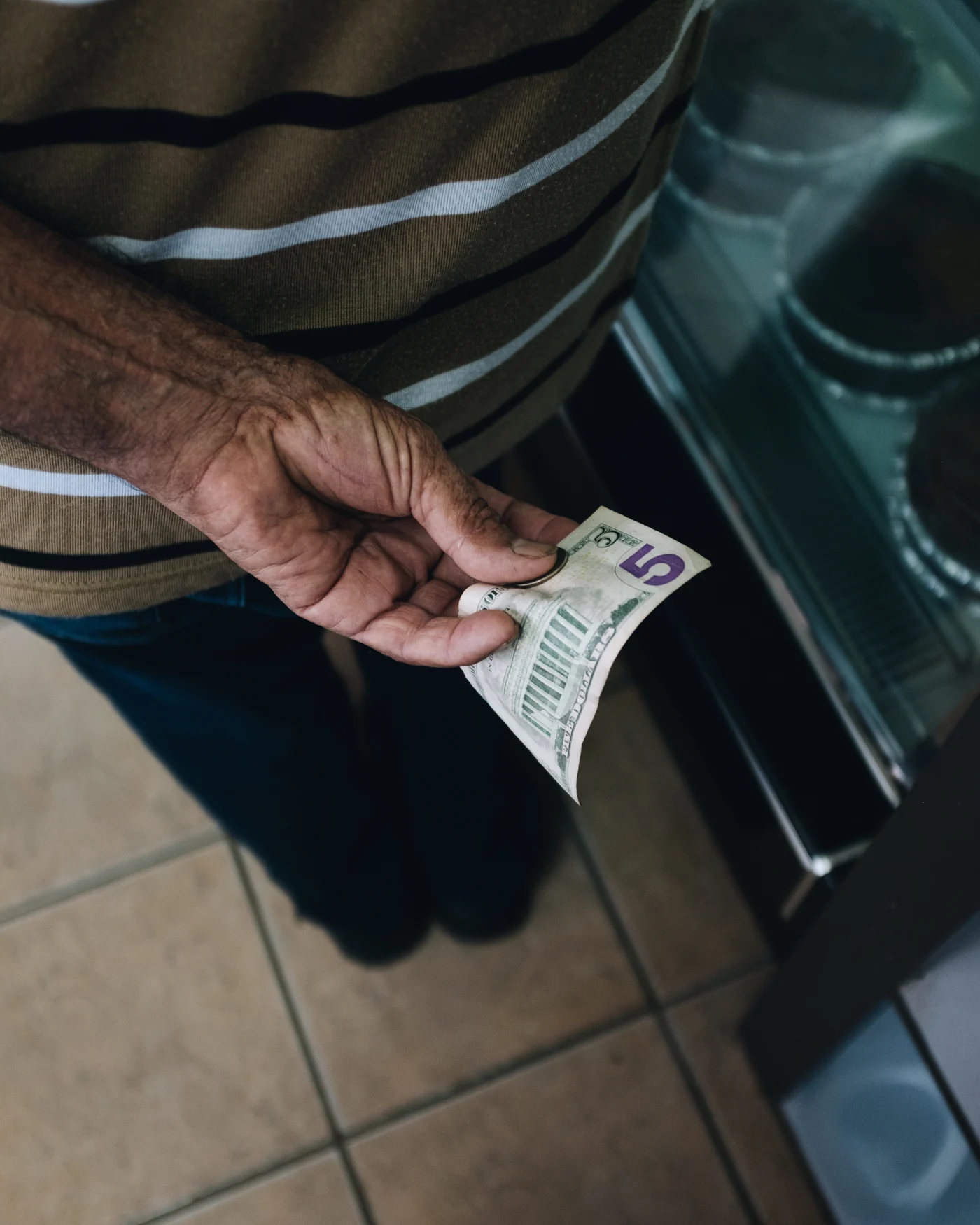 A weathered hand holding a folded five dollar bill at a bakery case
