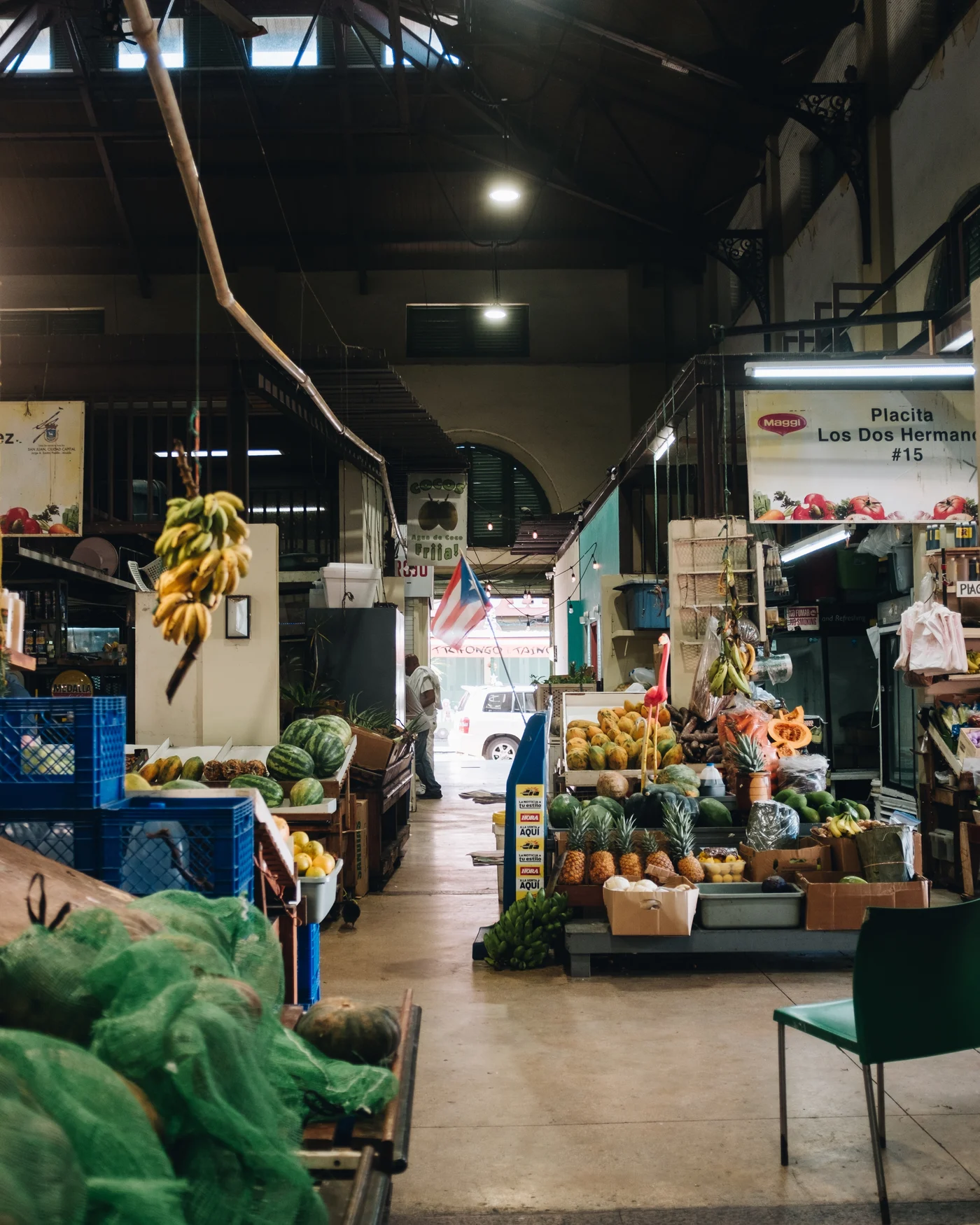 Inside a produce market, bananas hanging from a beam and a Puerto Rican flag over the aisle