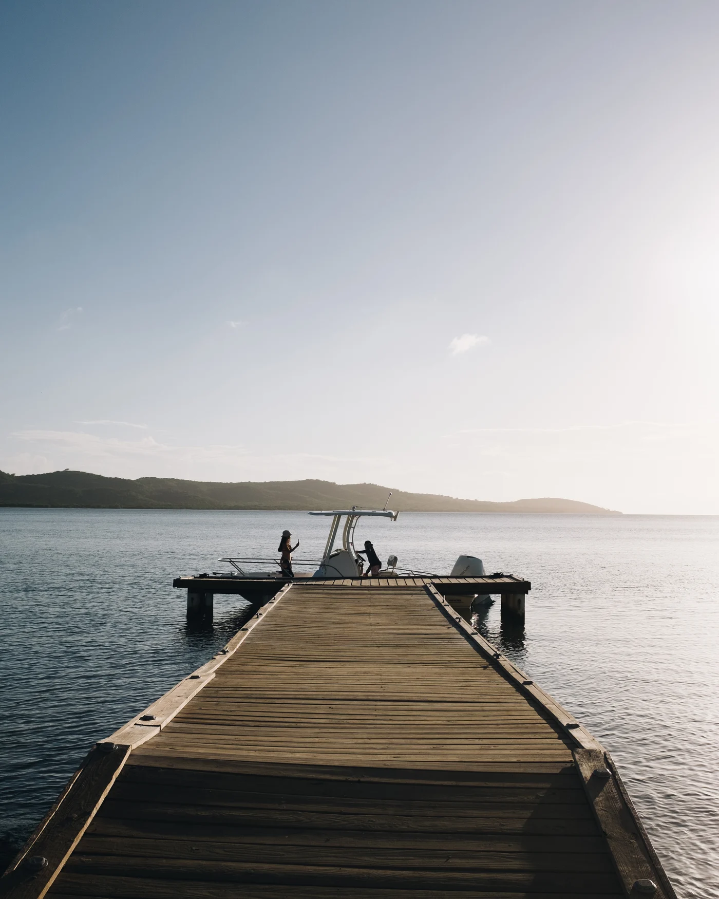 A long wooden dock running out to a moored boat, hills hazy across the water