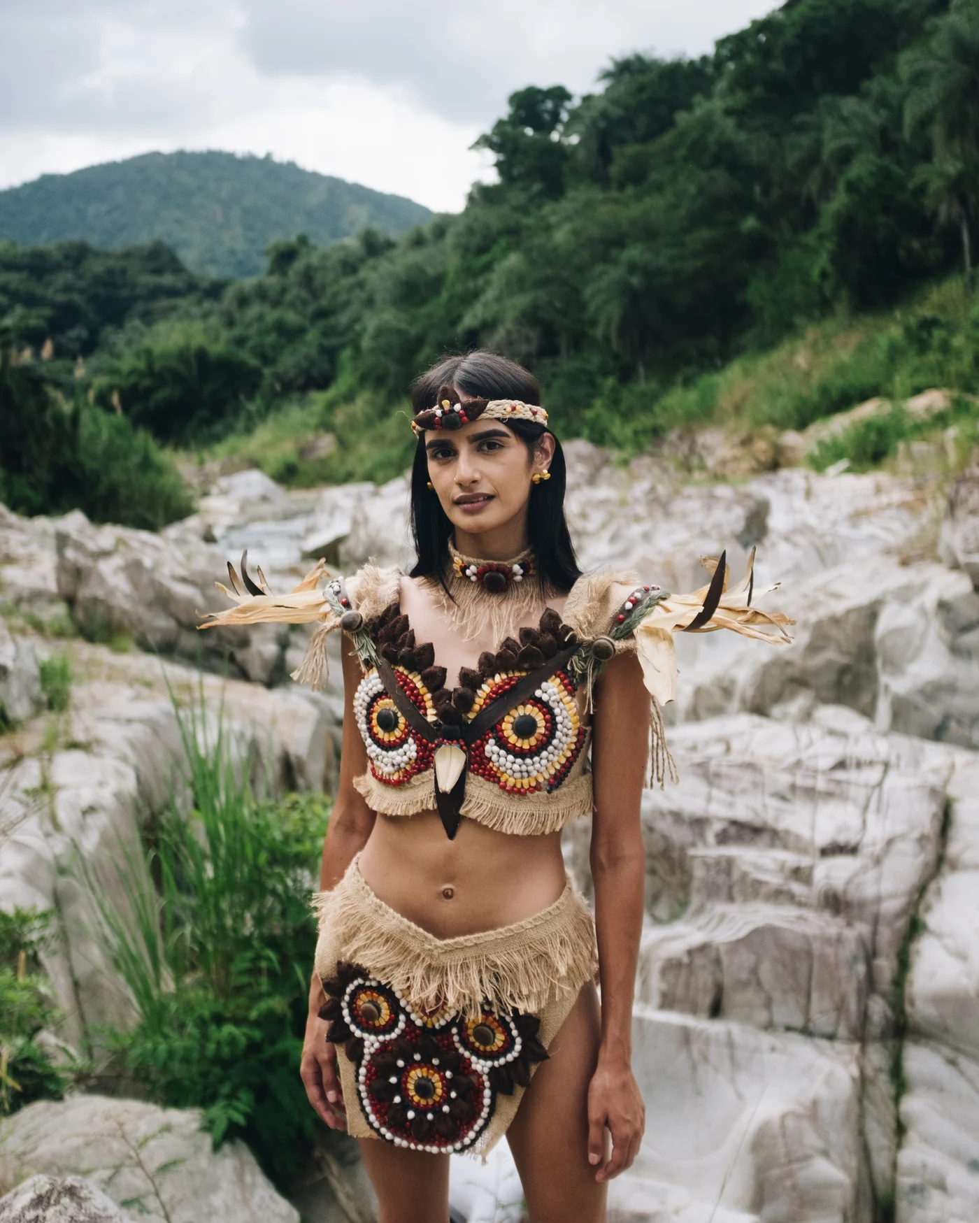 A model in a beaded owl costume standing among pale river boulders below green hills