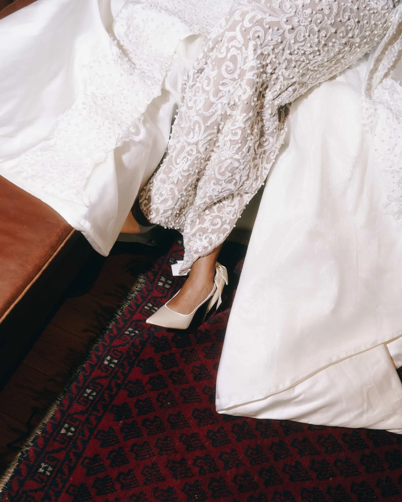 Looking down at a seated bride, the beaded lace sleeve and skirt of her gown spilling across a deep red patterned rug beside one satin heel