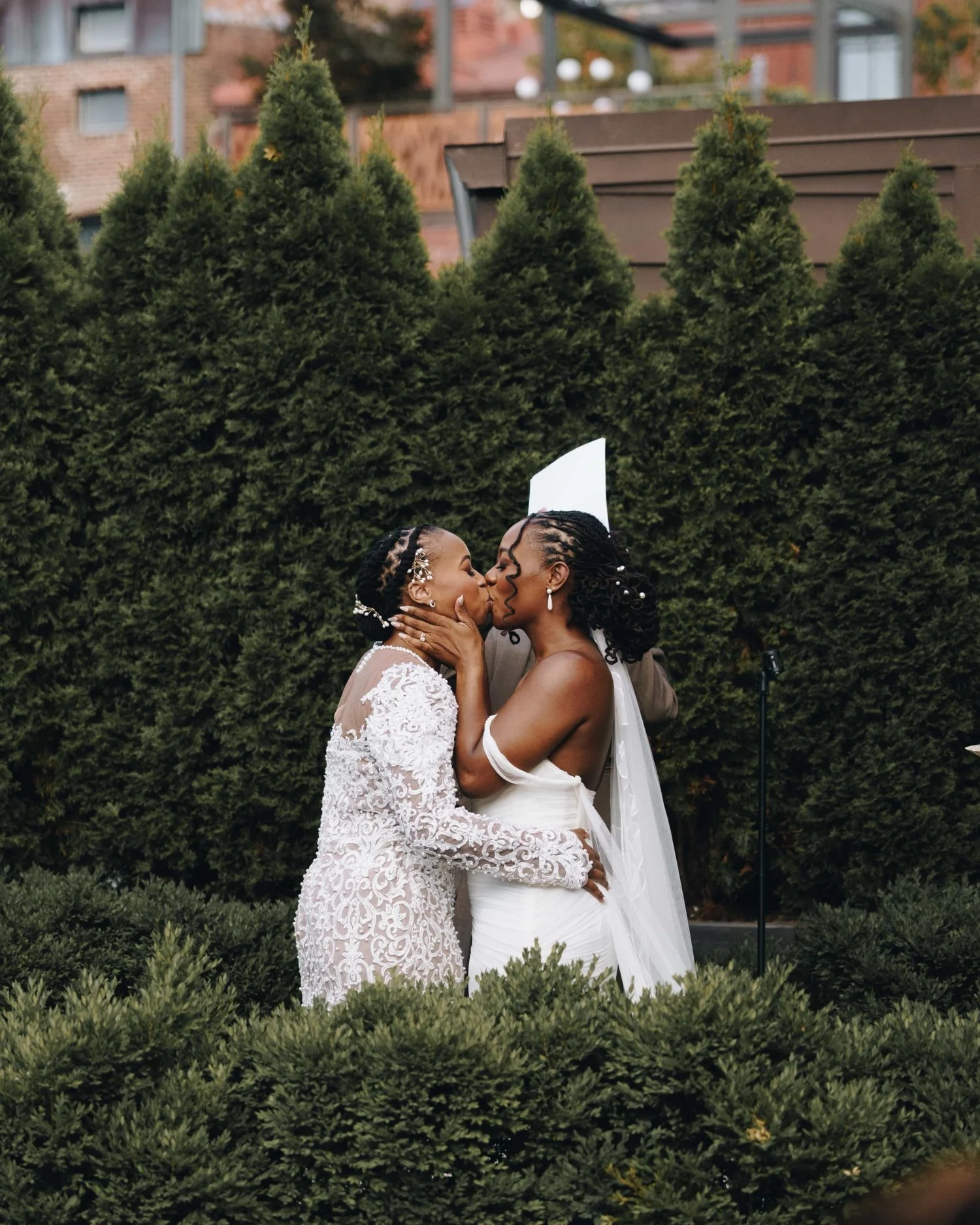 Two brides kissing in front of a wall of evergreens, one veil lifting behind them
