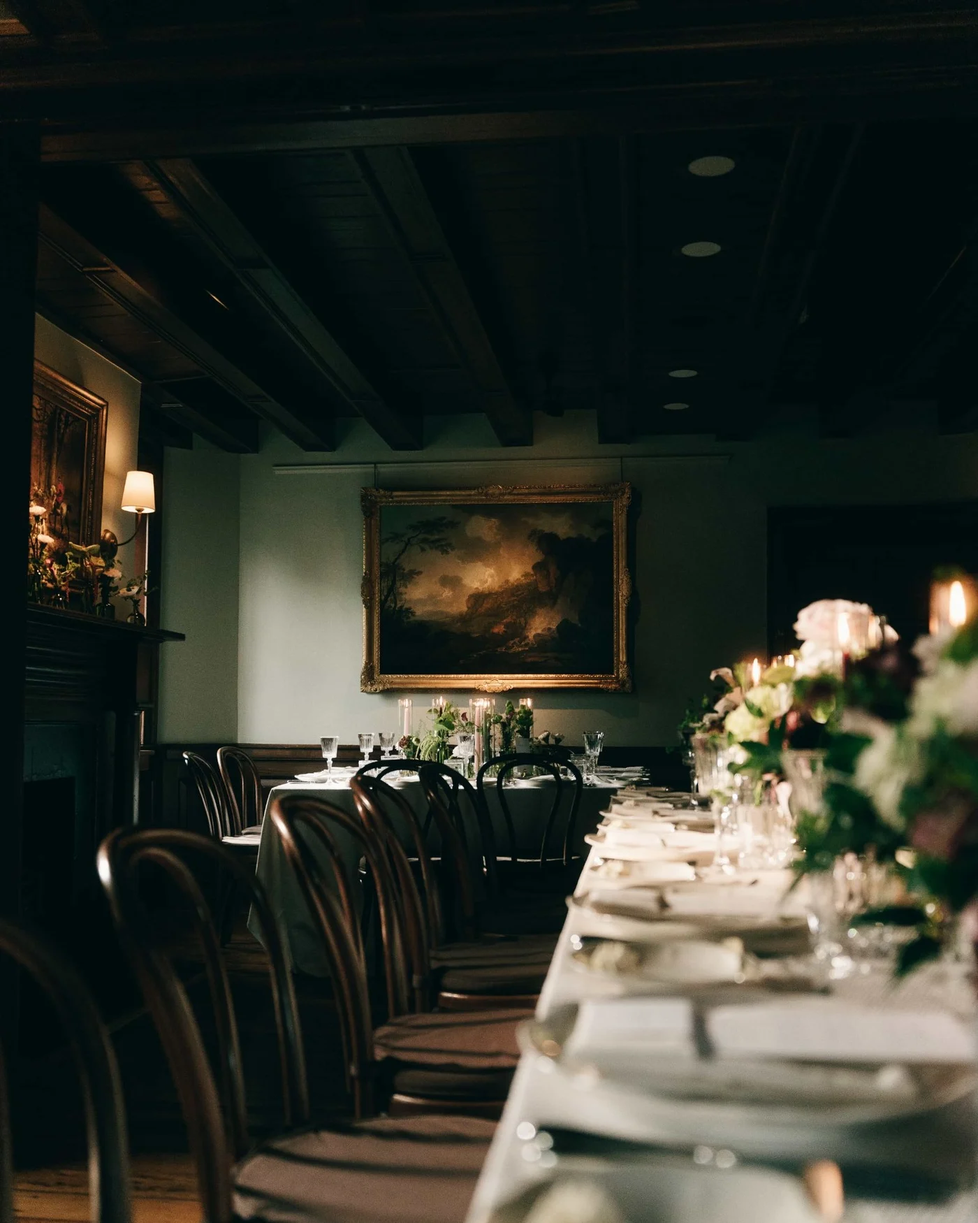 Another long table under a beamed ceiling, dressed with pale flowers, an oil painting hung above the fireplace