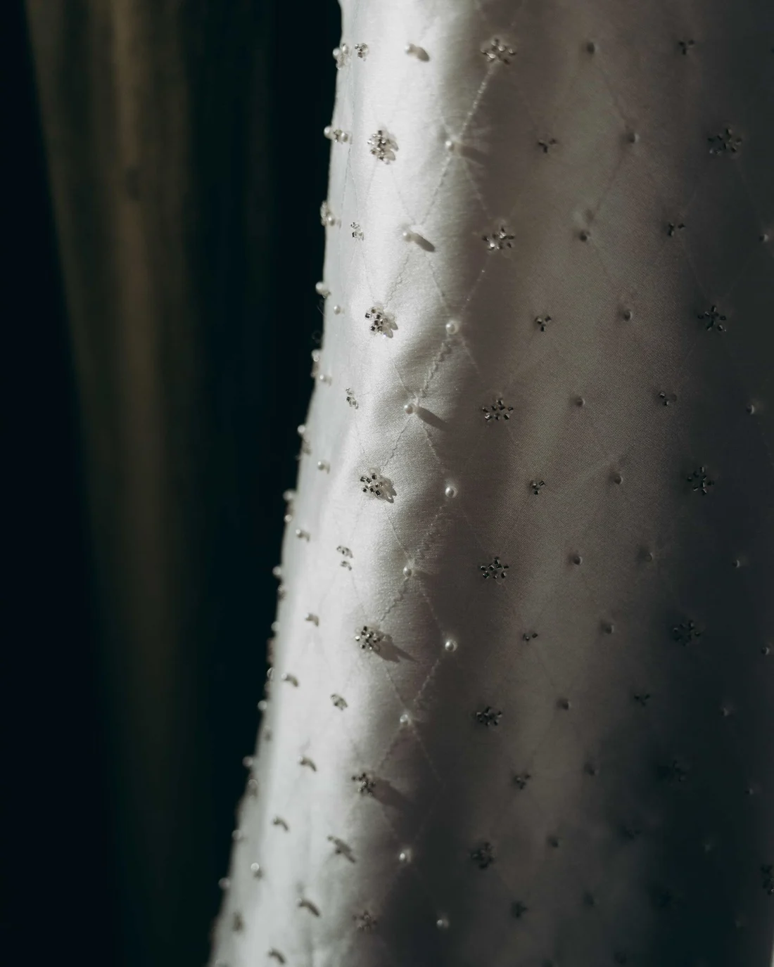 Close on a hanging gown, pearls and small crystal clusters scattered across quilted silk in a shaft of light