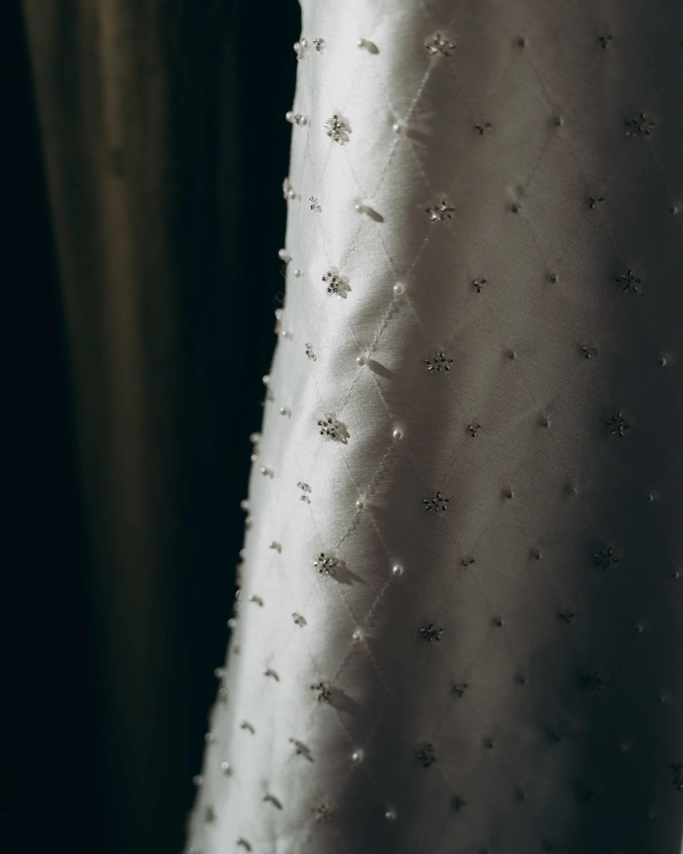 Close on a hanging gown, pearls and small crystal clusters scattered across quilted silk in a shaft of light