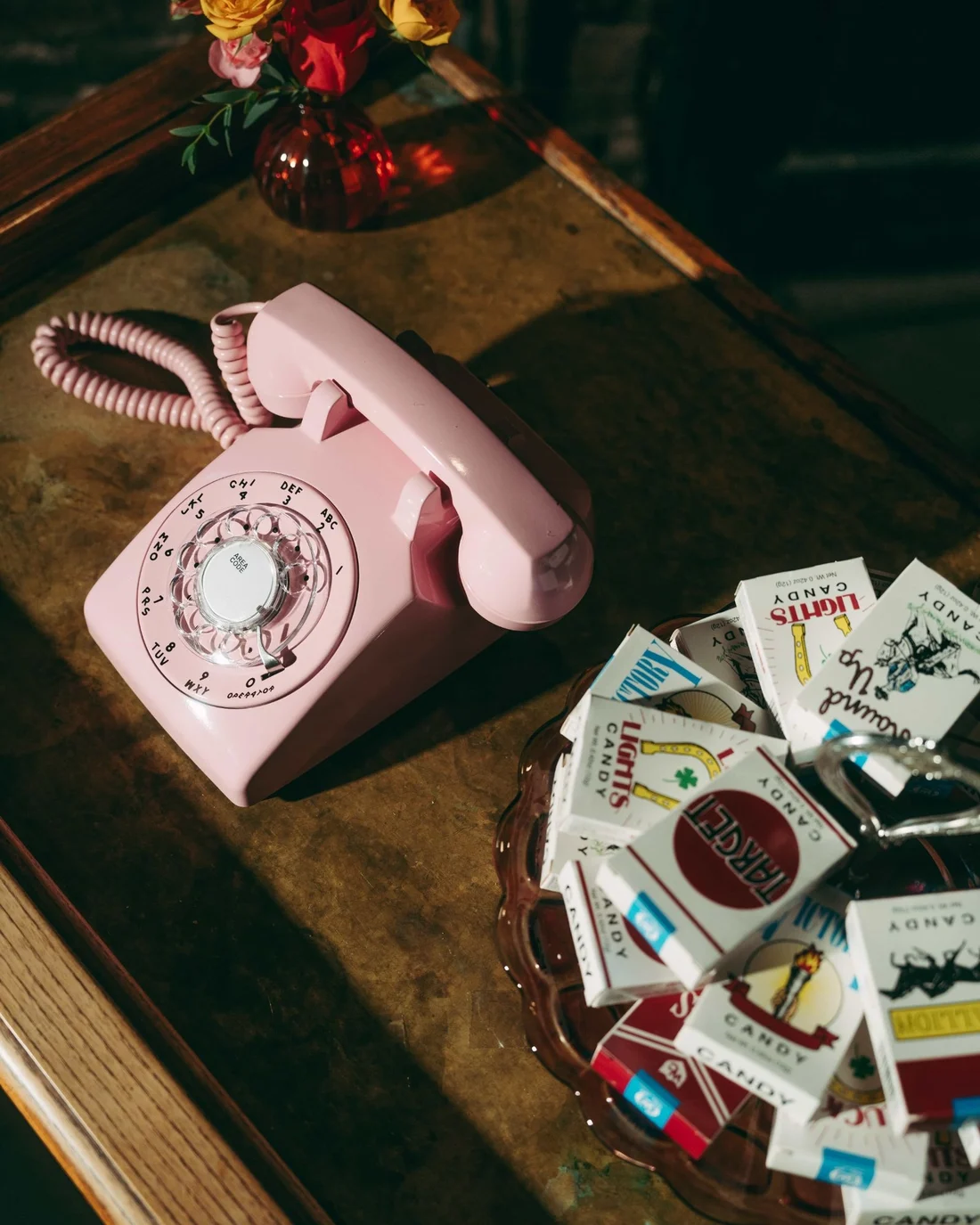 A pink rotary telephone on a worn table beside a dish heaped with candy cigarette packs