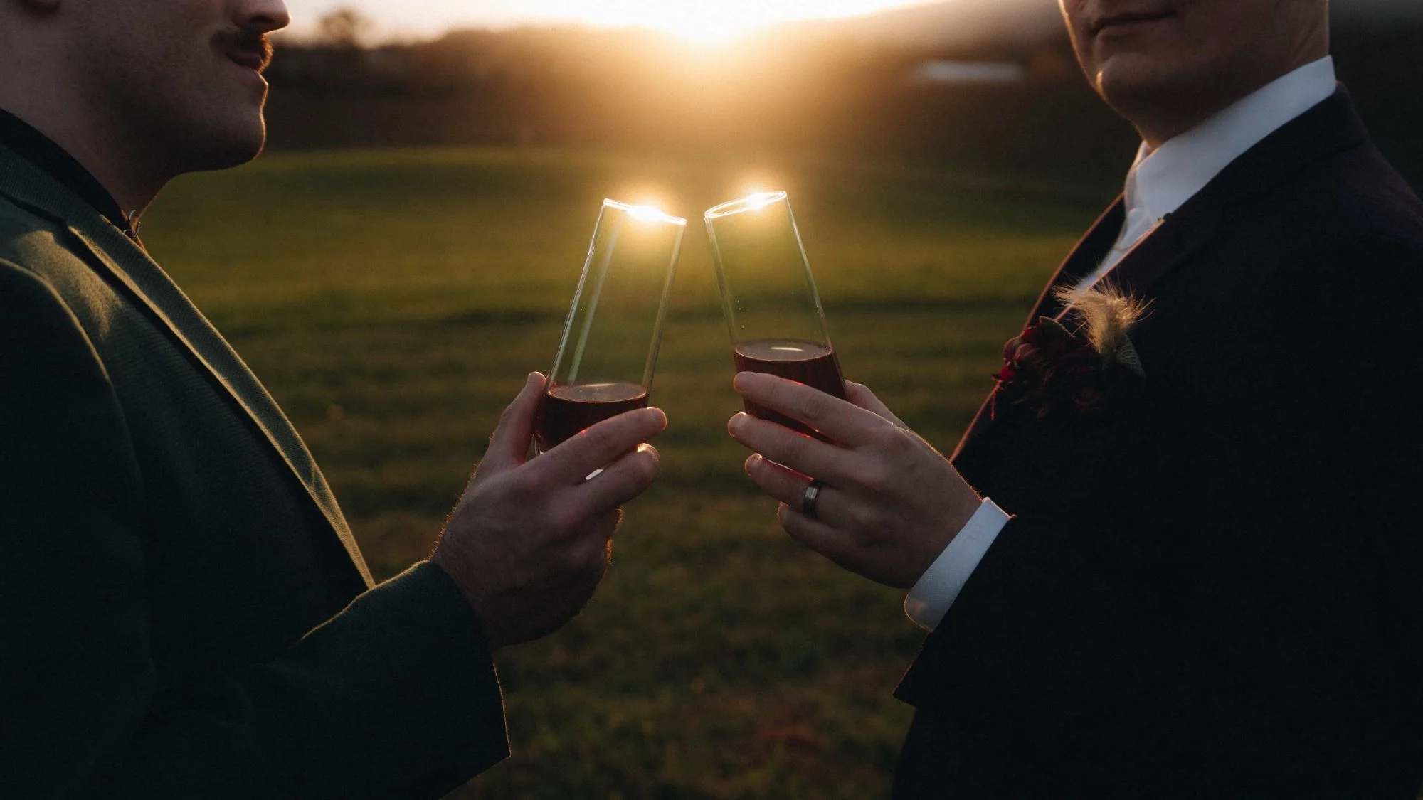 Two grooms touching glasses over an open field, the low sun burning through the wine