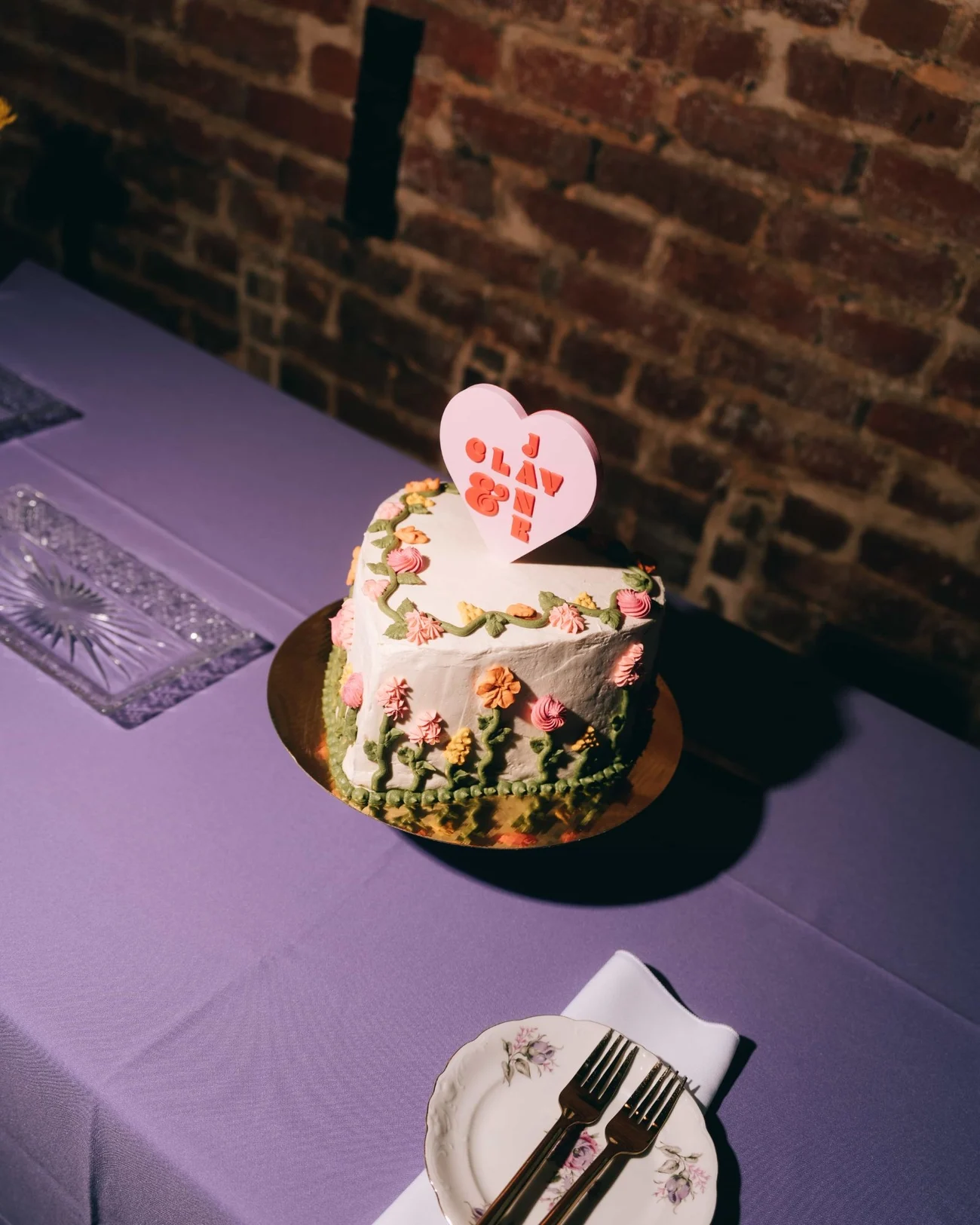A single tier cake iced with piped flowers under a heart shaped topper, on a violet cloth against exposed brick