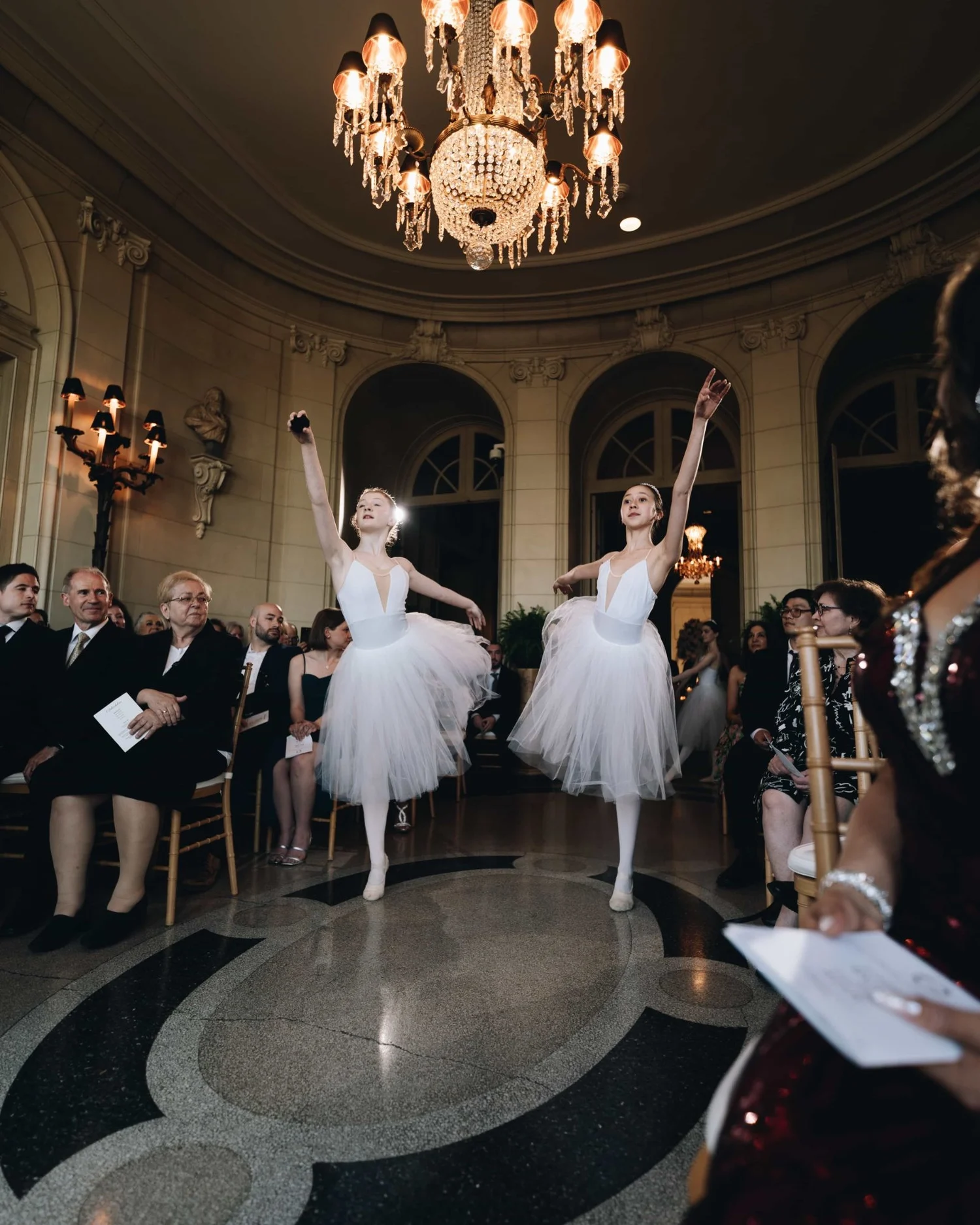 Two ballerinas dancing on a terrazzo floor beneath a chandelier while the guests watch from their seats