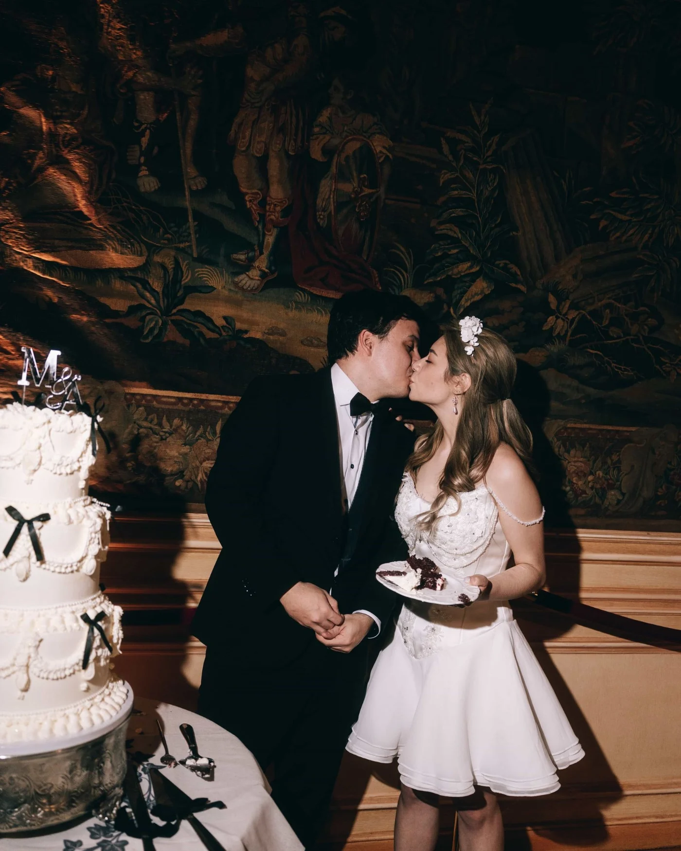 A couple kissing beside a tall tiered cake tied with black bows, a painted mural behind them