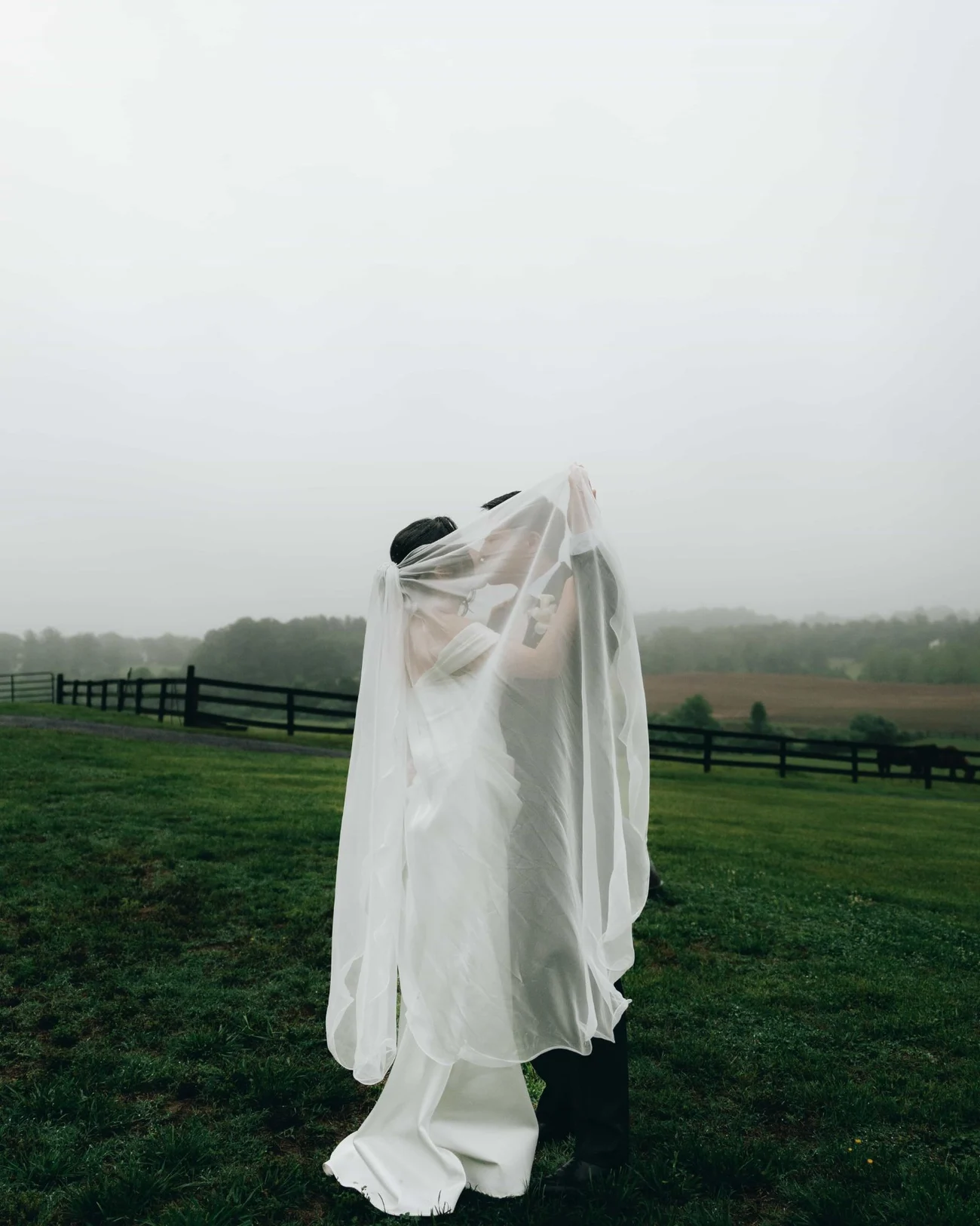 A bride holding her veil up over both of them, kissing the groom underneath it in a misty pasture