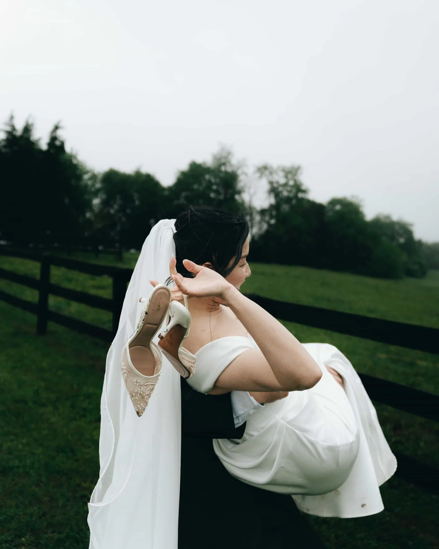 A groom carrying the bride across wet grass, her beaded heels dangling from her hand