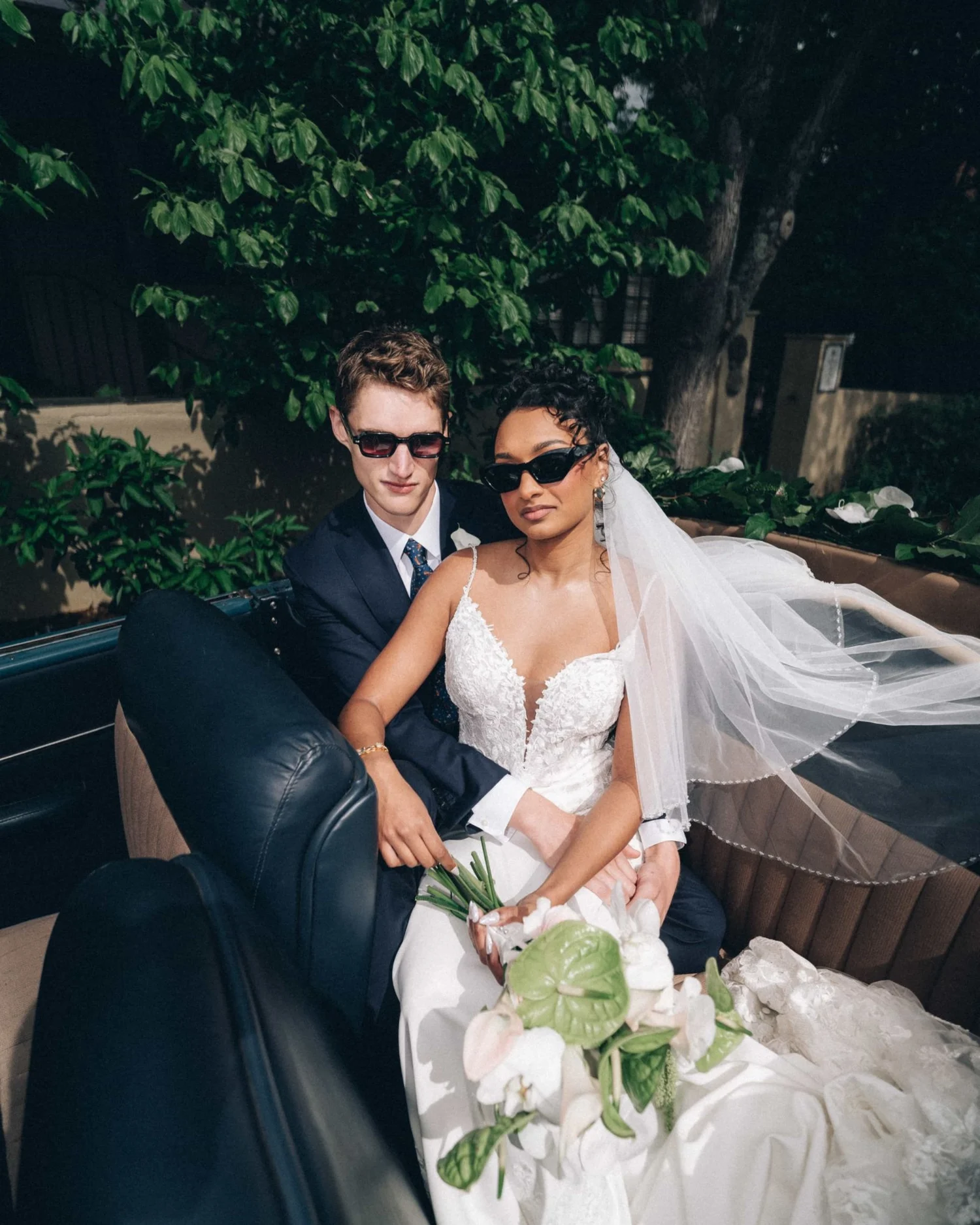 A couple in sunglasses riding in the back of an open car, the veil spilling over the door and lilies in her lap